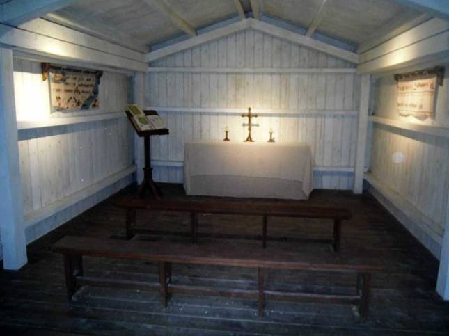 Small, wooden rectangular room with a small altar at the top and two benches to sit on.