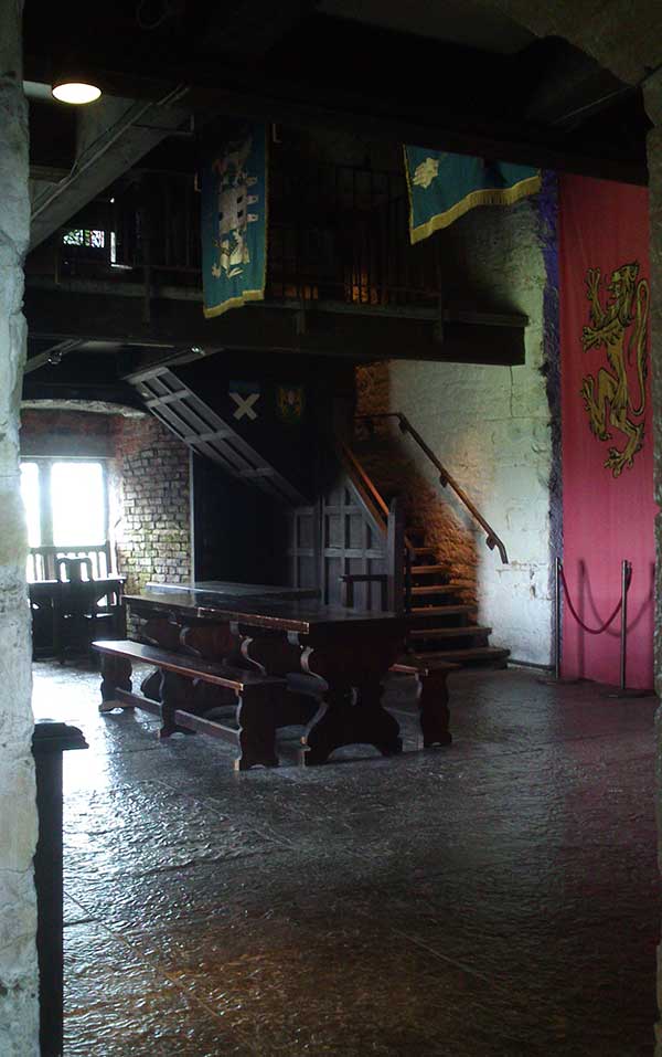 A large, dark, two-storey room with large table and chairs, wooden stairs to the next level and a large wall tapestry.