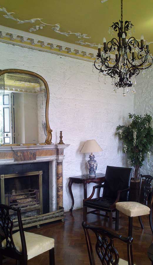 Chairs circling an ornate fireplace with large mirror above; also a beautiful chandelier hanging from a yellow ceiling.